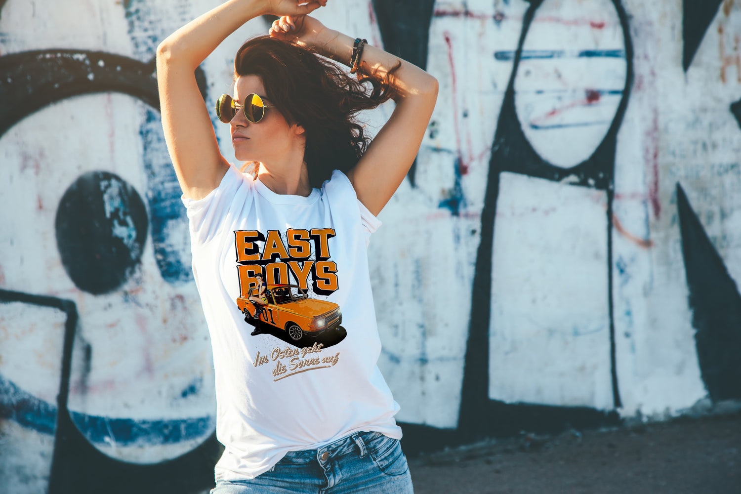 Frau mit weißem "East-Boys" T-Shirt posiert vor einer Graffiti-Mauer.